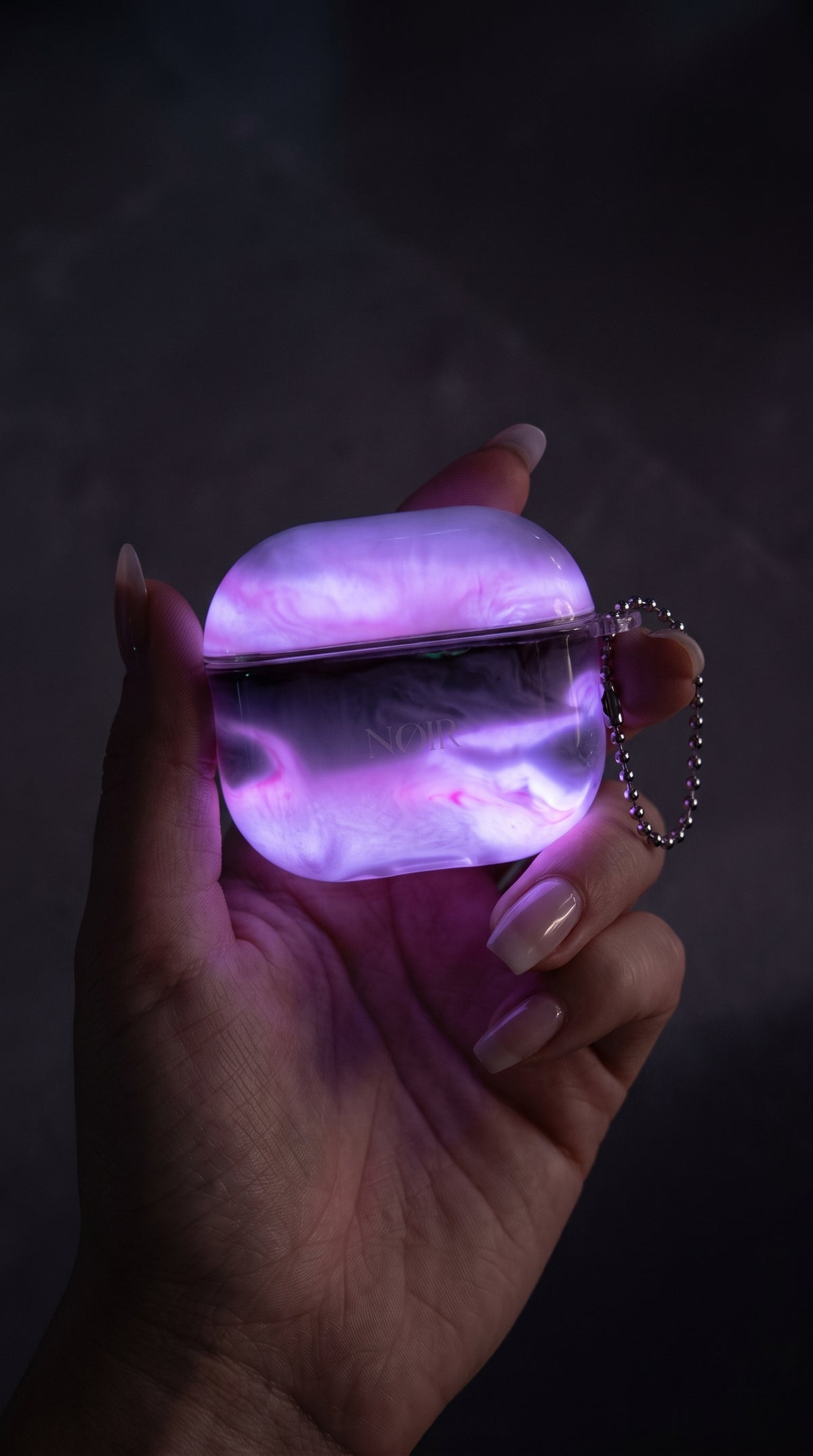 Realistic product photography: a glowing lavender and neon-pink marble AirPods protective case (matching the pink case in bottom-left of the reference screenshot) held in a hand in dark lighting. The