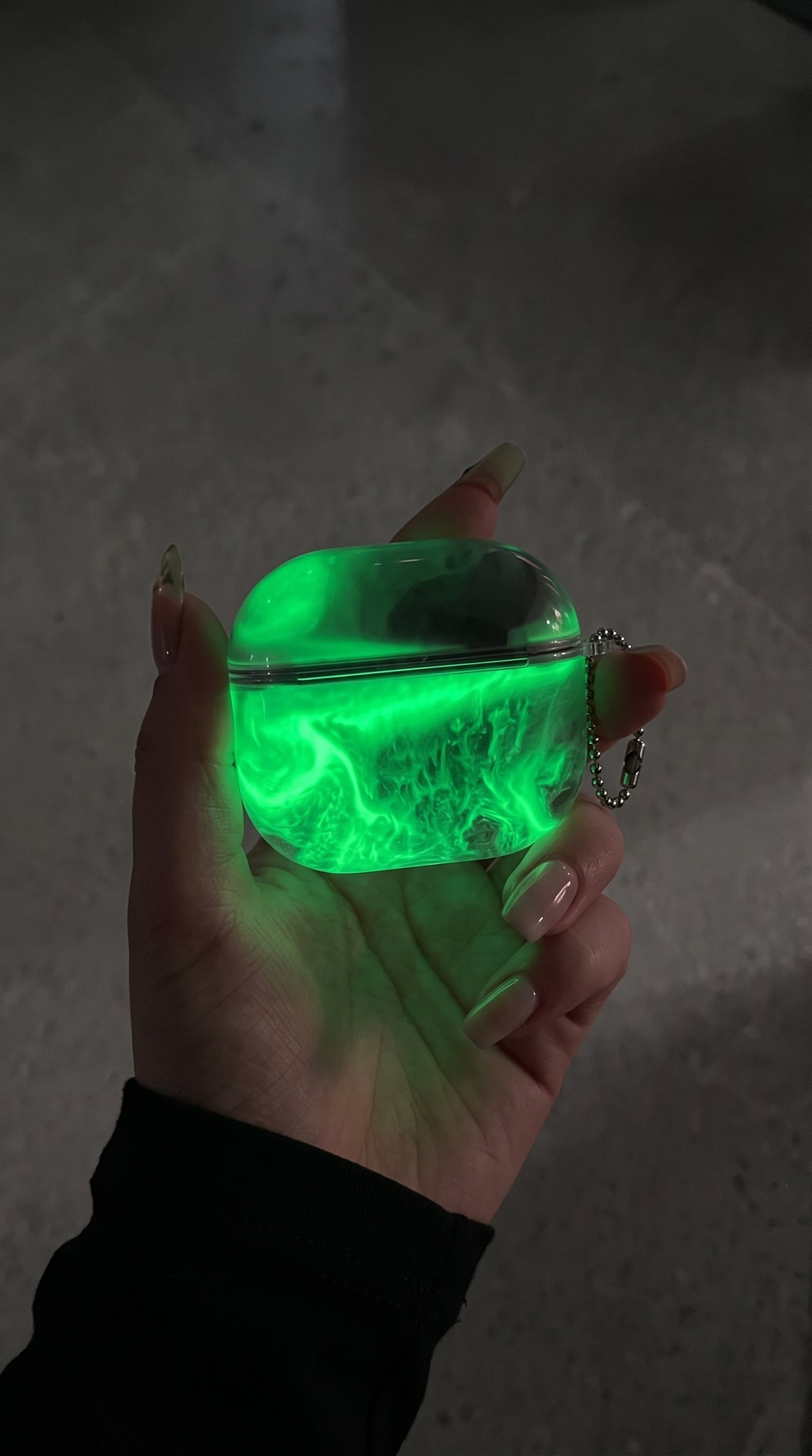 Realistic product photography: an electric emerald-green glowing marble AirPods protective case (matching the green case in the bottom-right of the reference screenshot) held in a hand in dim moody li